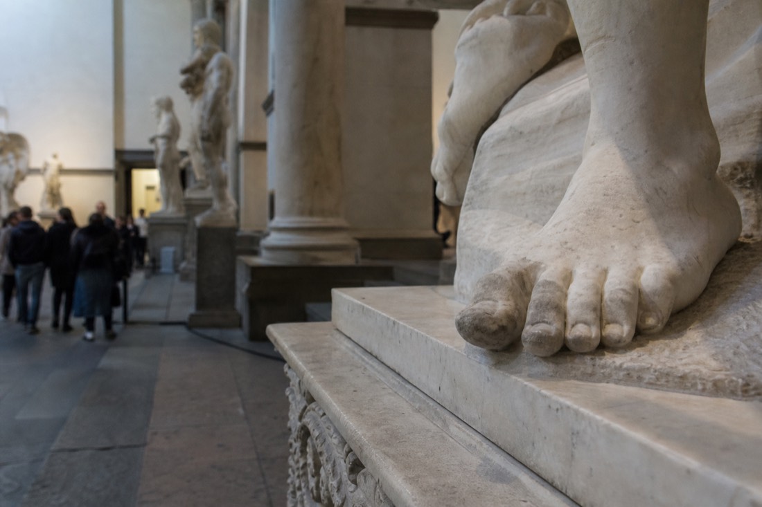 Close view of the marble left foot and toes of a monumental statue resting on a carved base under soft museum lighting