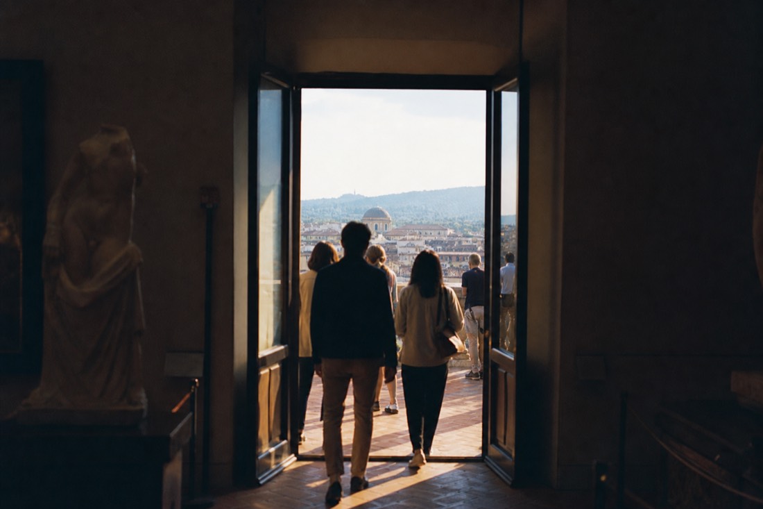 Silhouetted visitors seen from behind in the bright doorway of a Florence gallery with terracotta rooftops in the distance