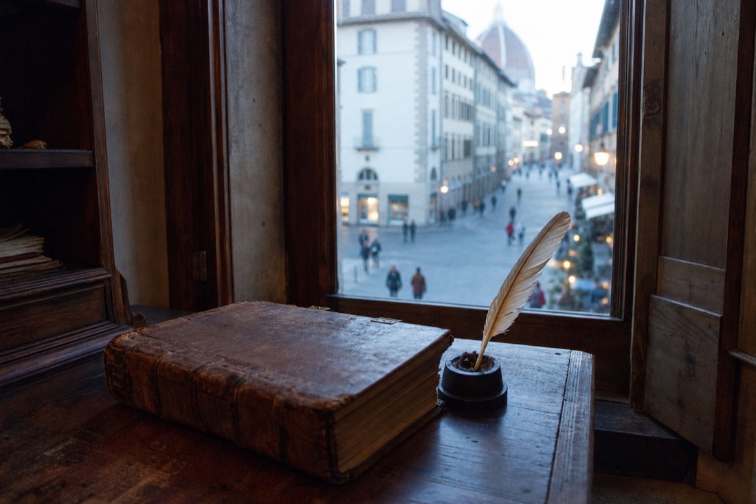 Old leather-bound ledger and a quill on a wooden desk beside a small window looking onto a Florence street, morning light