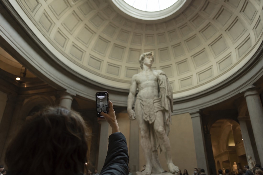 A phone screen held up in the Tribuna framing the full height of David, with the top of the statue near the domed skylight of the Accademia Gallery