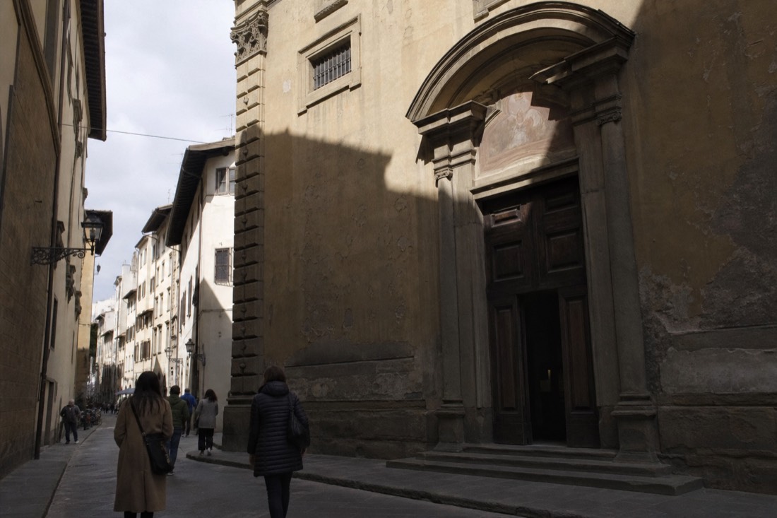 Narrow Florence side street with rough stone palazzo walls and deep afternoon shadow falling across a chapel doorway