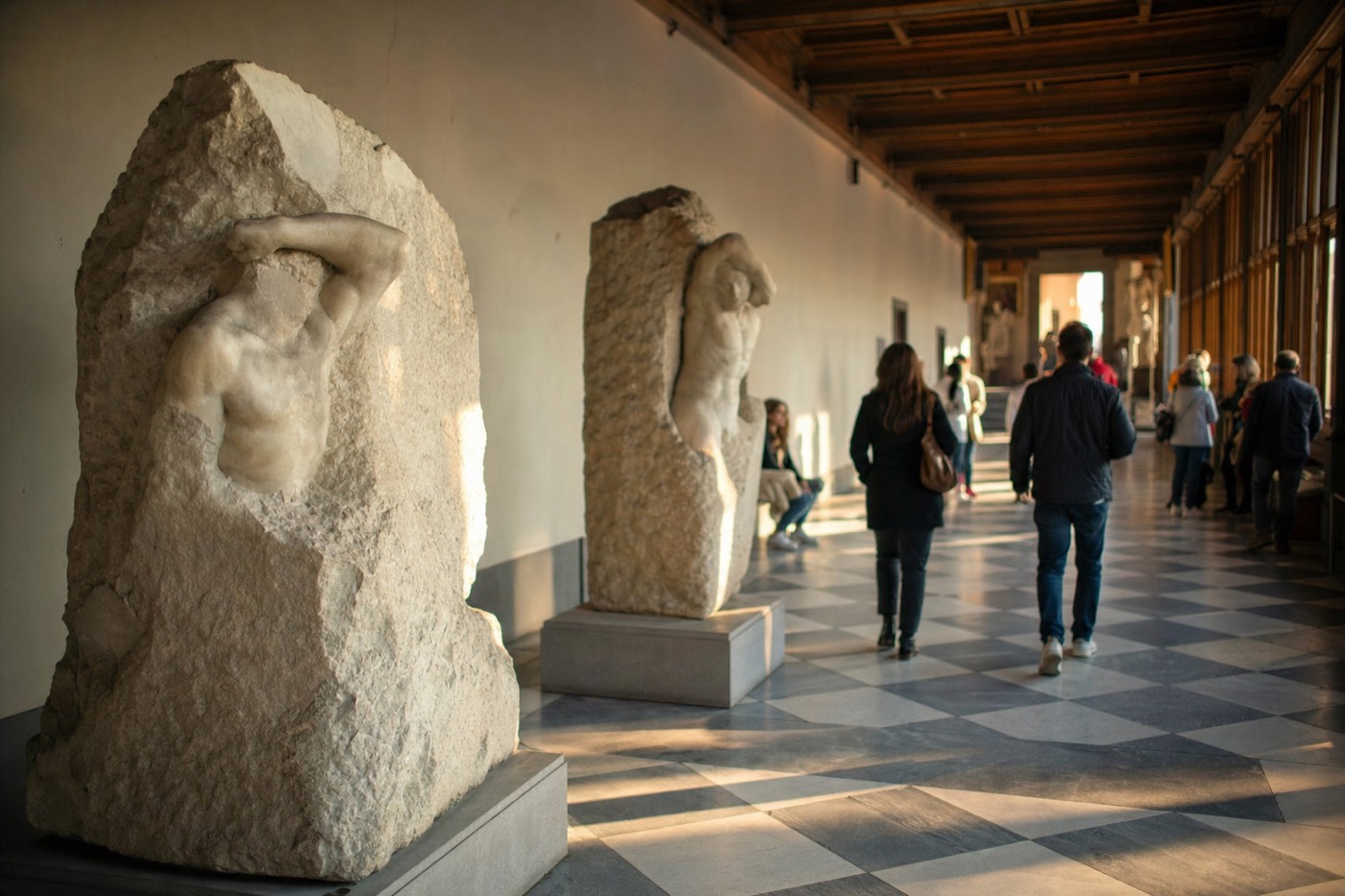 Four rough marble figures on plinths along a naturally lit gallery corridor in Florence, each partly carved out of its block