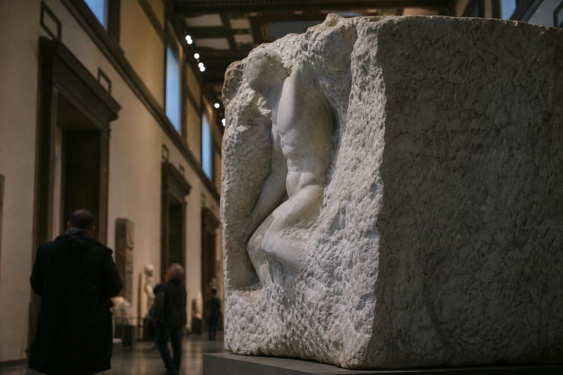 A large rough marble block on a museum plinth with a partly emerged human figure, dramatic overhead gallery light in Florence