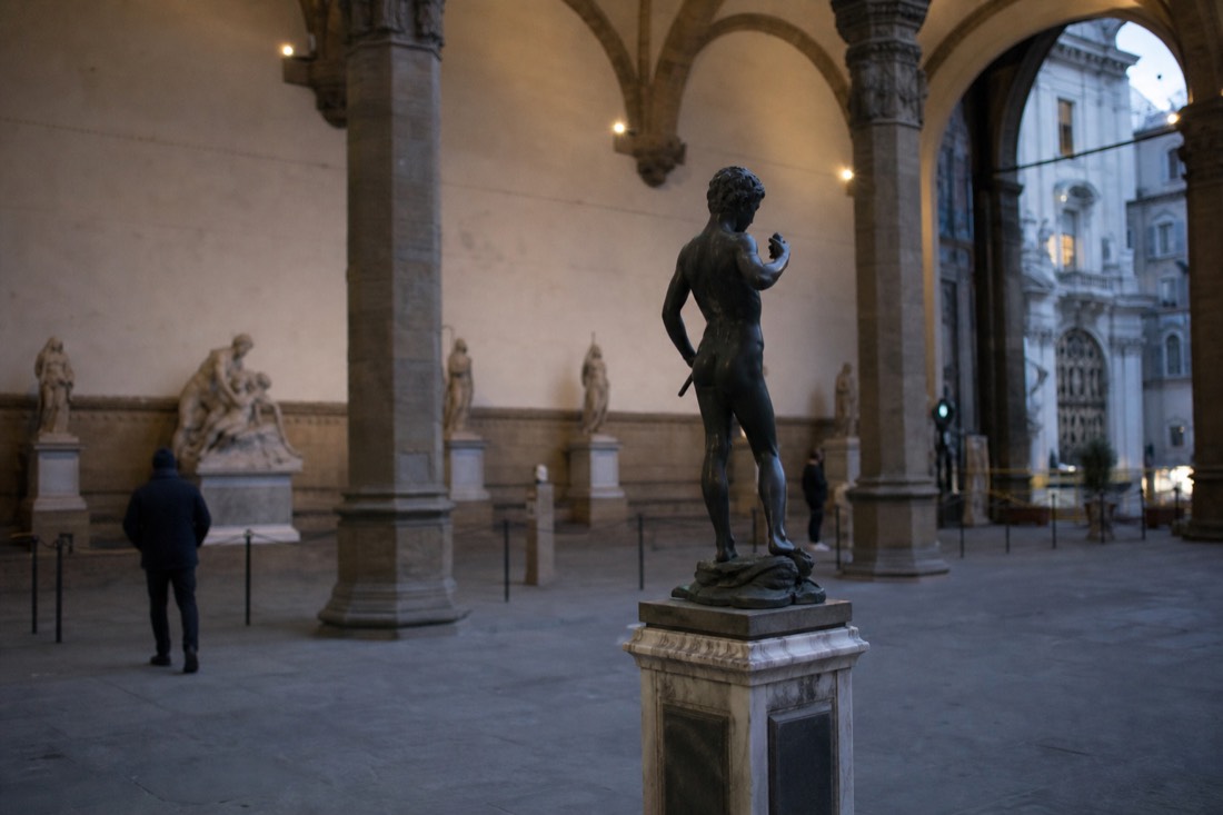 A small dark bronze statue of a youth on a pedestal in a stone-vaulted museum hall in Florence