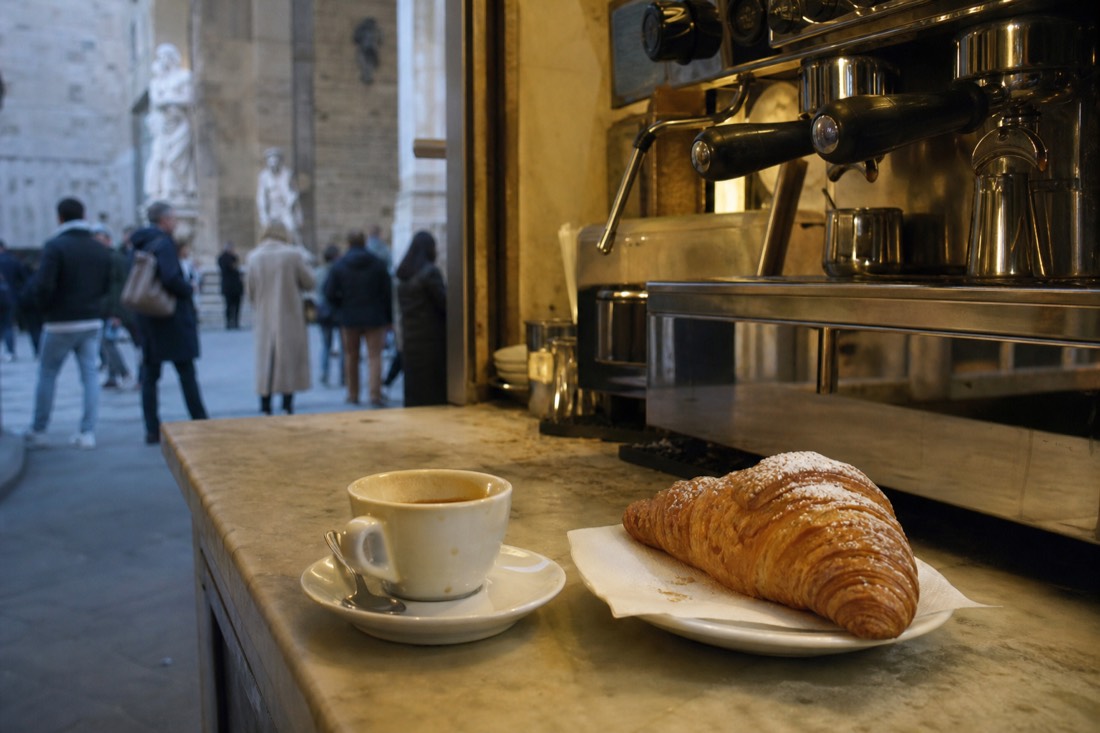 A single espresso and a cornetto on a marble bar counter with a chrome espresso machine behind, early morning light near the Accademia