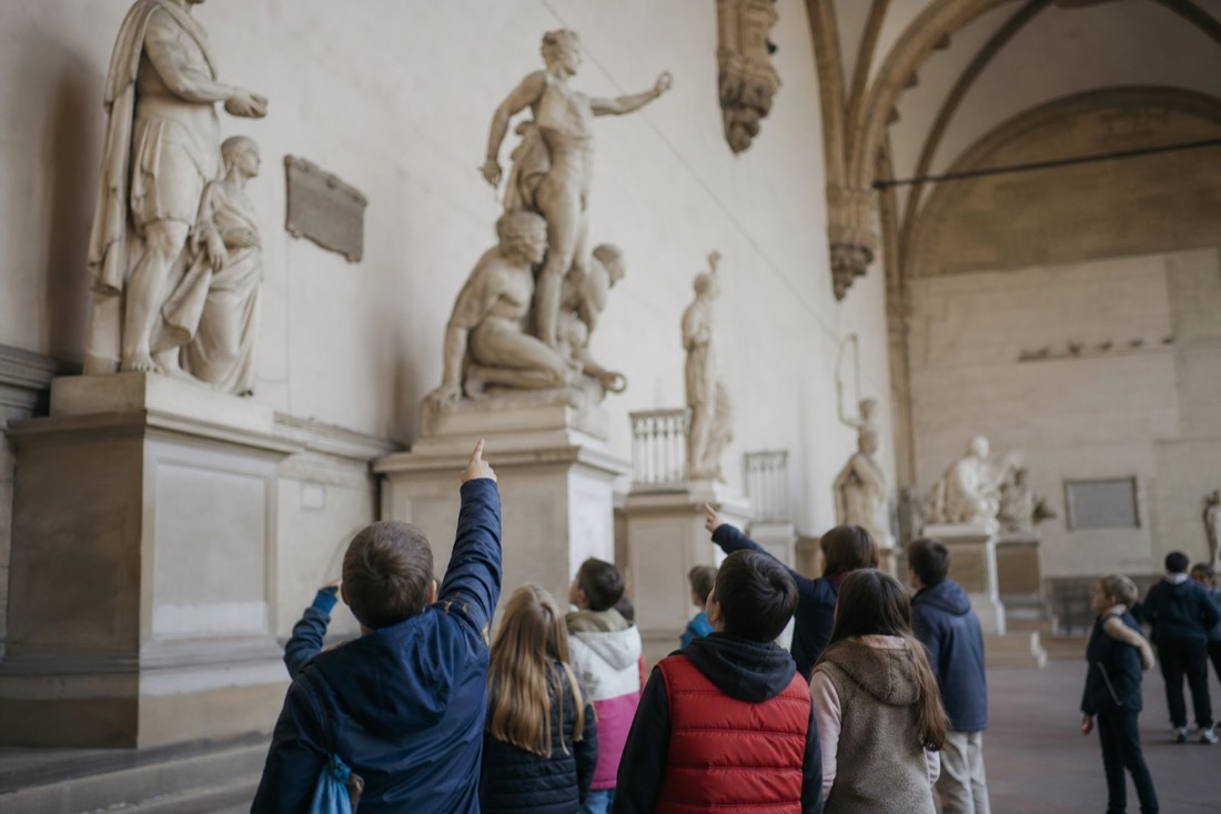 Young children pointing up at large marble statues inside the bright sculpture hall of the Accademia Gallery in Florence