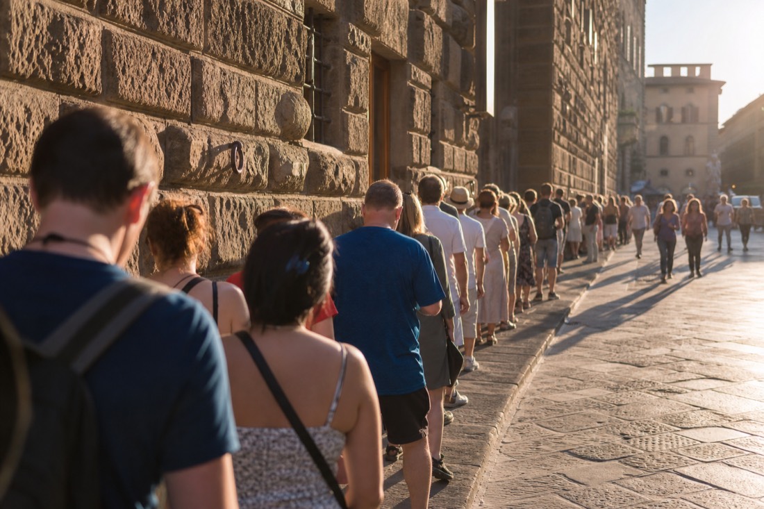 A line of tourists waiting in bright direct sunlight on a shadeless Florence street beside a tall stone building