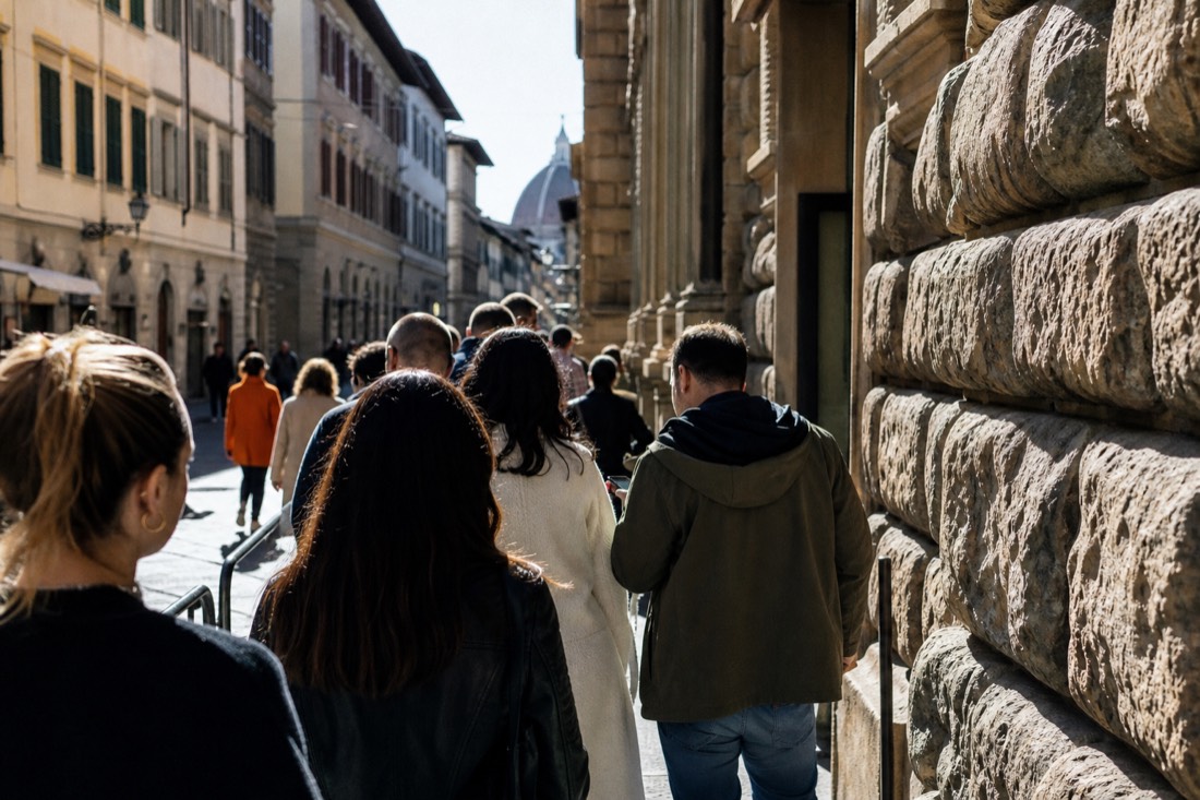 Visitors queued along a sunlit Florence street beside the stone wall of a gallery on a busy morning