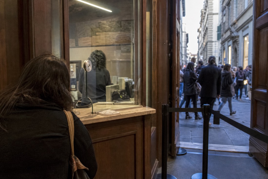 A small ticket pickup office on Via Ricasoli in Florence with paper tickets on the counter and the museum street visible through the doorway