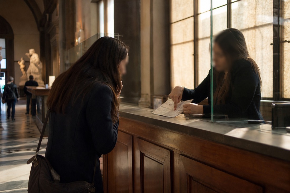 A visitor looking surprised at the ticket counter of the Accademia Gallery while staff point to a printed price list