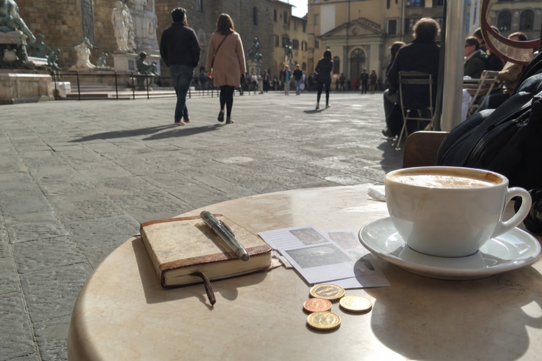 A small paper notebook on a Florence cafe table with two museum tickets and a few euro coins beside a cappuccino
